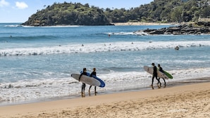 Una playa con surfistas caminando por la orilla, un litoral rocoso y una colina con edificios al fondo.