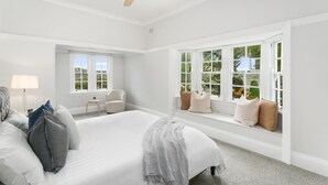 Un dormitorio con una cama grande, un ventilador de techo, un asiento junto a la ventana con cojines y una mesita de noche con lámpara.