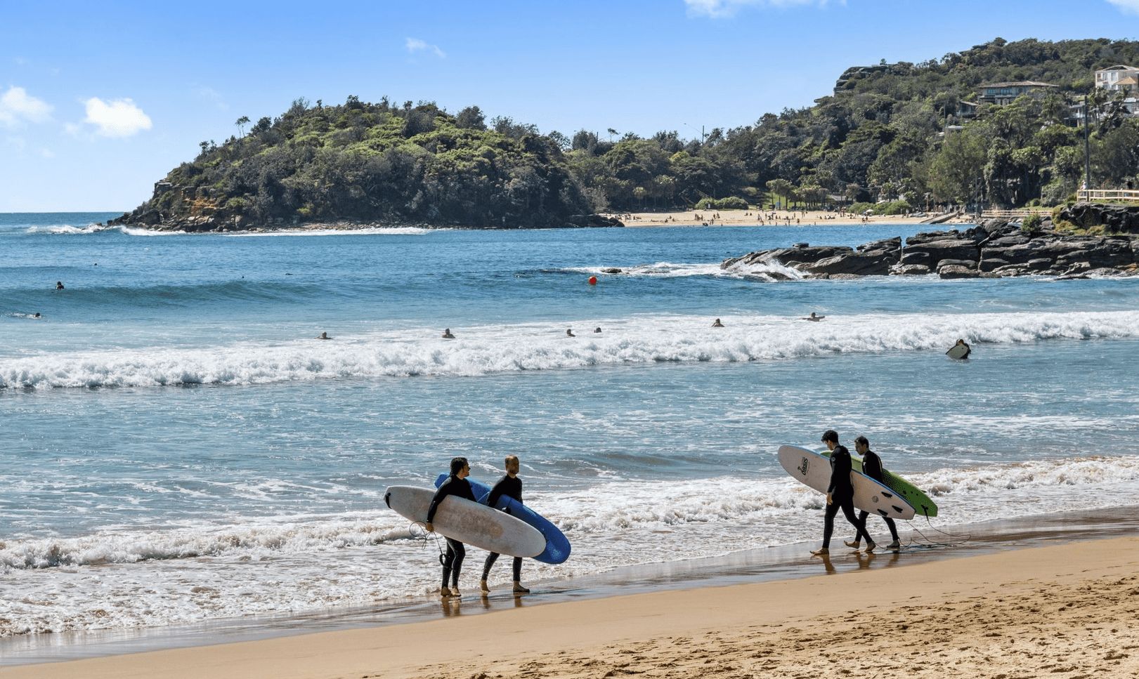 Une plage où l’on voit des surfeurs se promener le long du rivage, une côte rocheuse et une colline avec des bâtiments en arrière-plan.