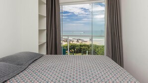 A bedroom with a bed, a window with curtains, and a view of the beach. - Apartamento Espetacular em Bombinhas (Bombinhas)