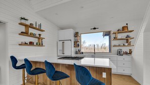 Una cocina moderna con armarios blancos, una isla central con taburetes de bar, estantes de madera con utensilios de cocina y un amplio ventanal con vistas a árboles.