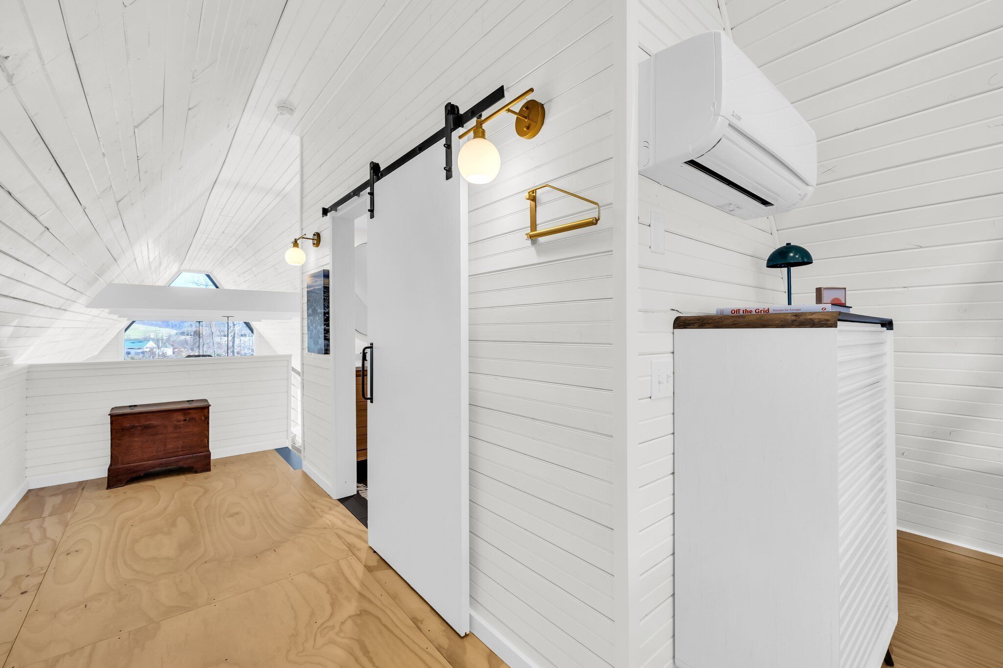 A room with white wooden walls, a wooden floor, a sliding door, a small wooden chest, a wall-mounted air conditioner, and a lamp on a shelf.