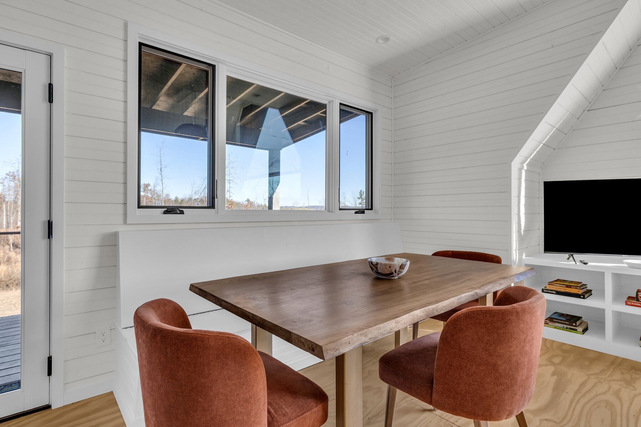 Un comedor con mesa de madera y cuatro sillas, ventanas amplias y un televisor sobre un estante blanco.