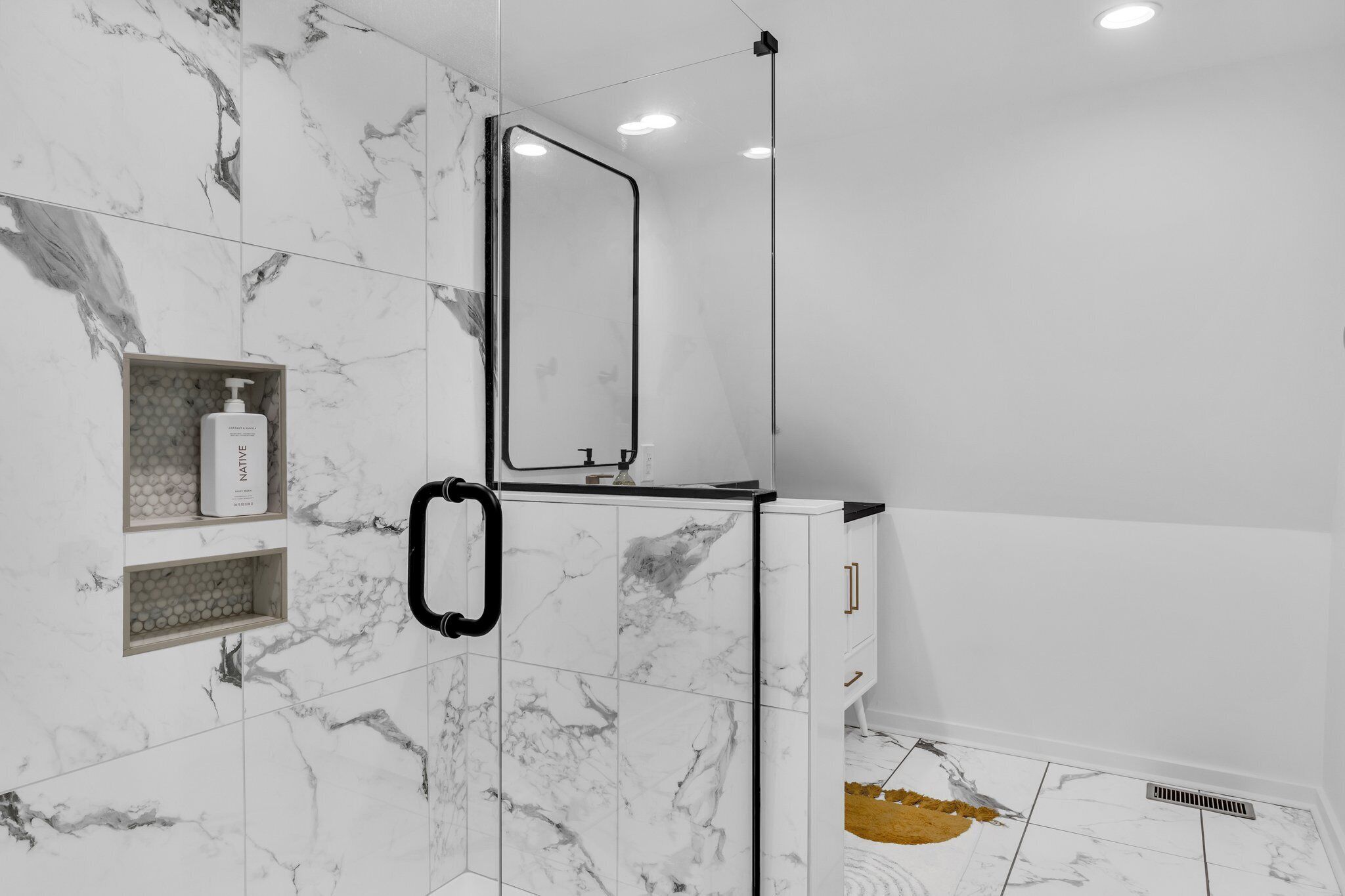 A bathroom with marble tiles, a mirror, a shower enclosure, and a soap dispenser.