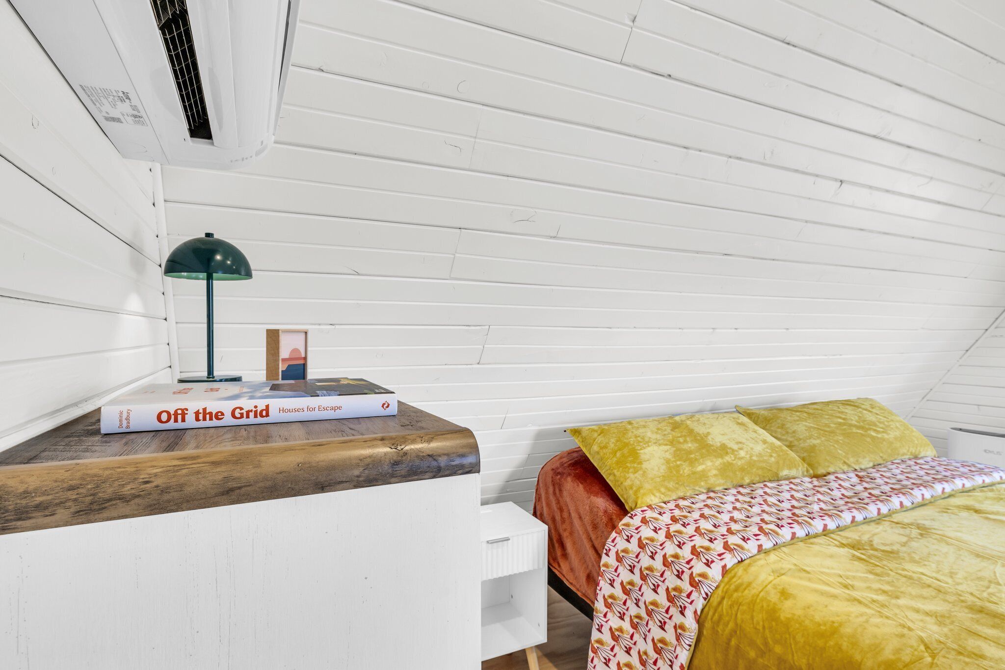 Un dormitorio con una cama, una mesita de noche, una lámpara y un libro titulado Casas fuera de la red para escapar.
