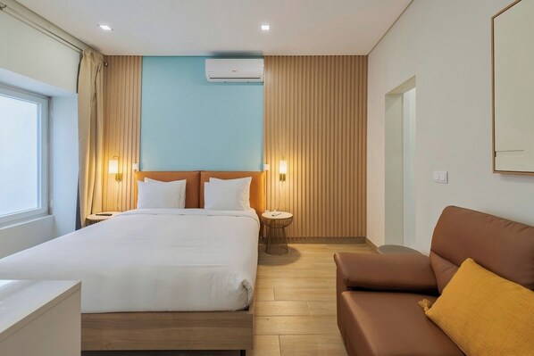 A hotel room with a bed, a brown sofa, a round side table, and a wall-mounted air conditioner. - Canto Doce Troino (Setúbal)