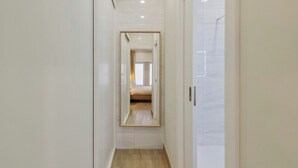 A narrow hallway with a wooden floor and white walls, leading to a bedroom with a bed and a window. - Canto Doce Troino (Setúbal)