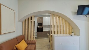 A modern living room with a sofa, a TV mounted on the wall, and a kitchen area visible through an arched doorway. - Canto Doce Troino (Setúbal)
