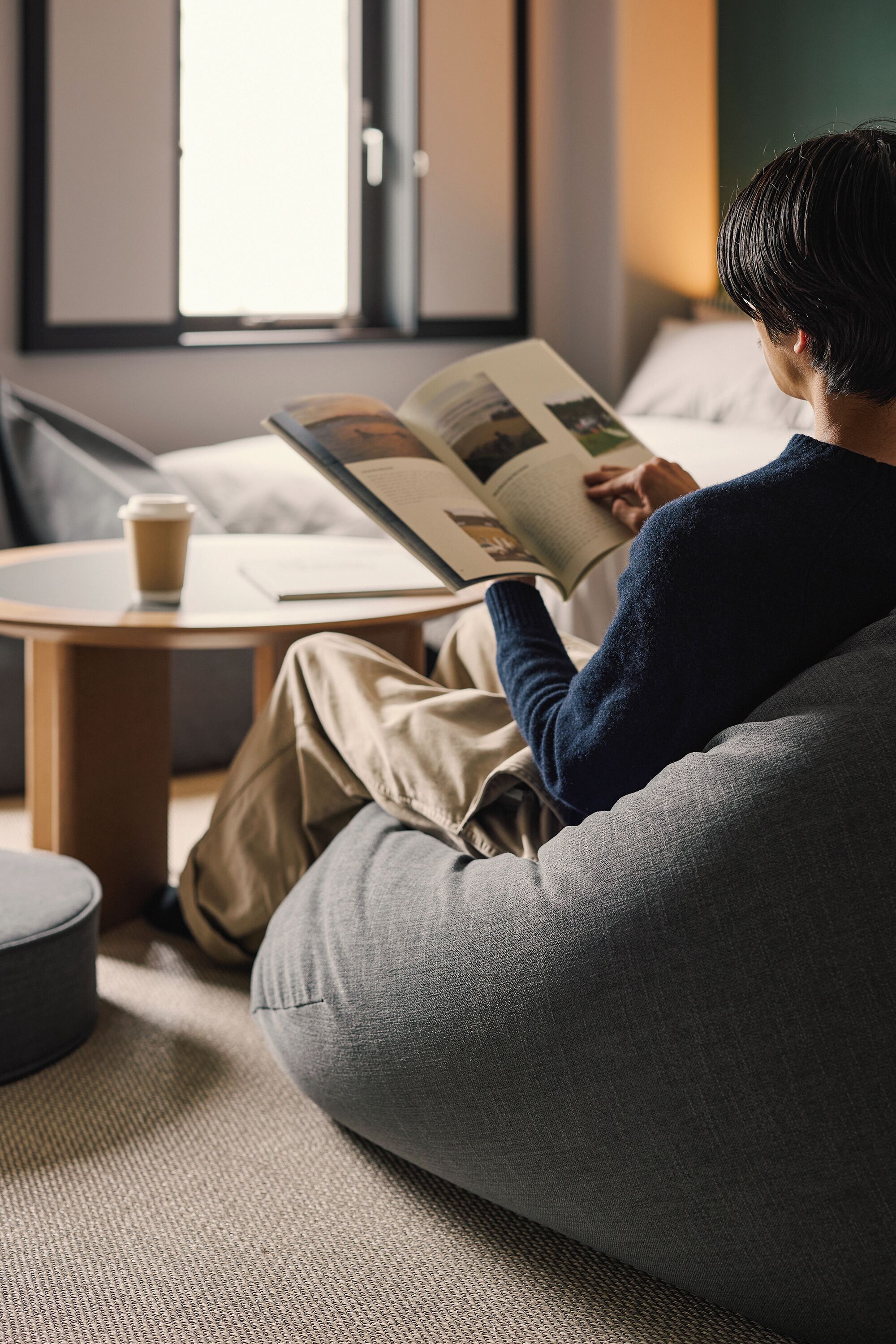 A person sitting on a grey bean bag chair reading a book with a coffee cup on a small round table beside them.