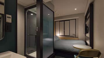 A modern hotel room with a glass-enclosed shower, a bed with a wooden headboard, a small round table, and a chair.