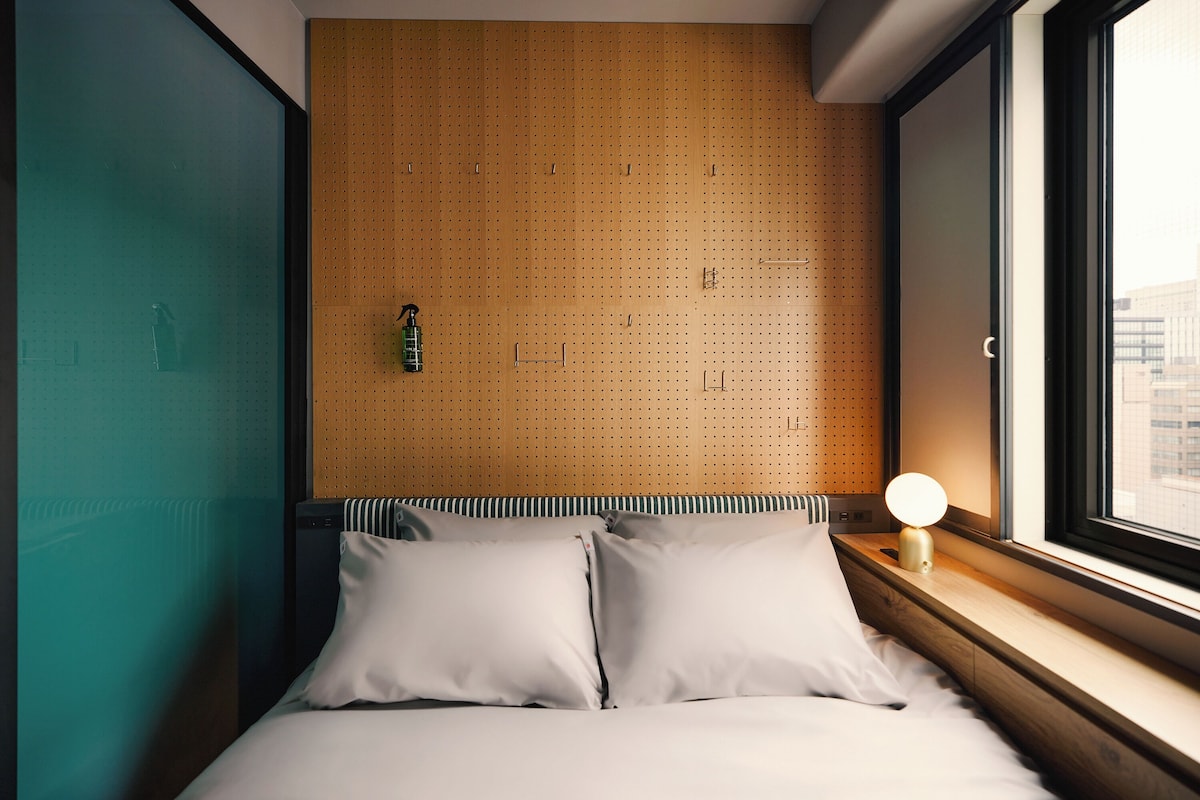 Een net opgemaakt bed met witte lakens en twee kussens, een wand van houten prikbord en een nachtkastje met een lamp.