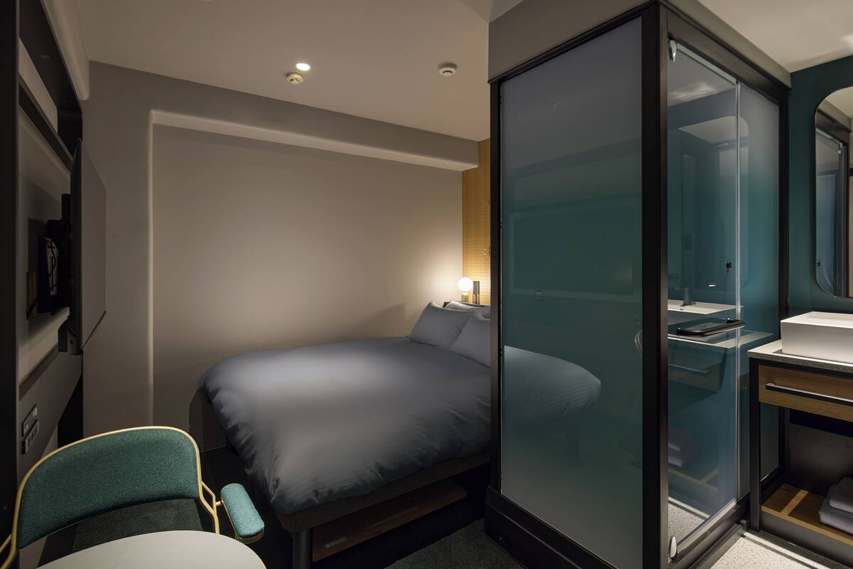 Een compacte hotelkamer met een bed, een klein bureau, een stoel, een glazen scheidingswand en een badkamer met spiegel en wastafel.
