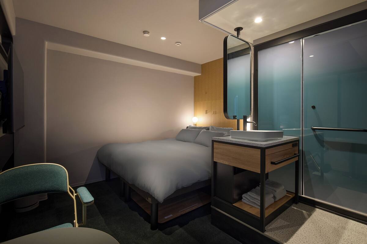Een moderne hotelkamer met een bed, nachtkastje met lamp, stoel en een garderobe met spiegel.