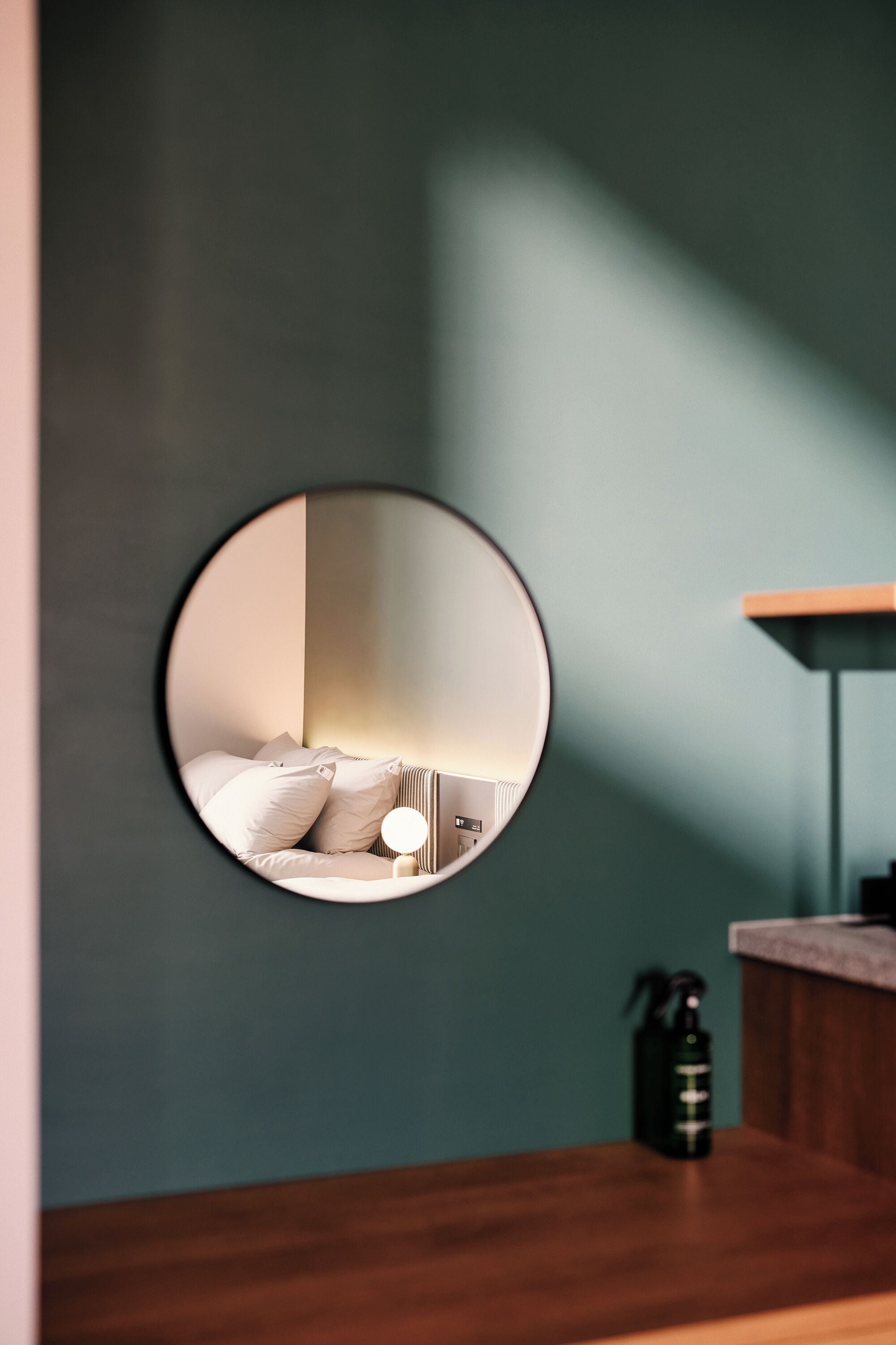 A round mirror reflecting a bed with pillows and a bedside lamp, a wooden counter, and a fire extinguisher on a teal wall.
