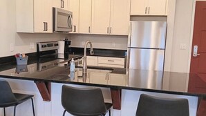 Une cuisine moderne avec des armoires blanches, des appareils en acier inoxydable et un comptoir noir. On y trouve trois tabourets de bar et des luminaires suspendus au-dessus de l’îlot central.
