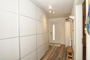 Een gang met witte betegelde muren, een houten vloer, een deur met een spiegel en een klein houten krukje.
