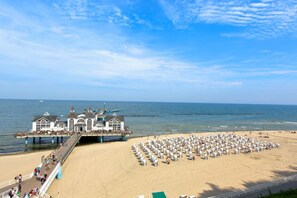 Een pier aan het strand met een rij strandstoelen en parasols, een gebouw met balkon, en de oceaan op de achtergrond.