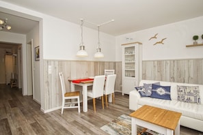 A modern living room with a white sofa, wooden coffee table, and a dining area with a red tablecloth. - Walidyll Residence, "Kleiner Stubber" Vacation Rental - with pool access at AHOI Sellin - (Sellin)