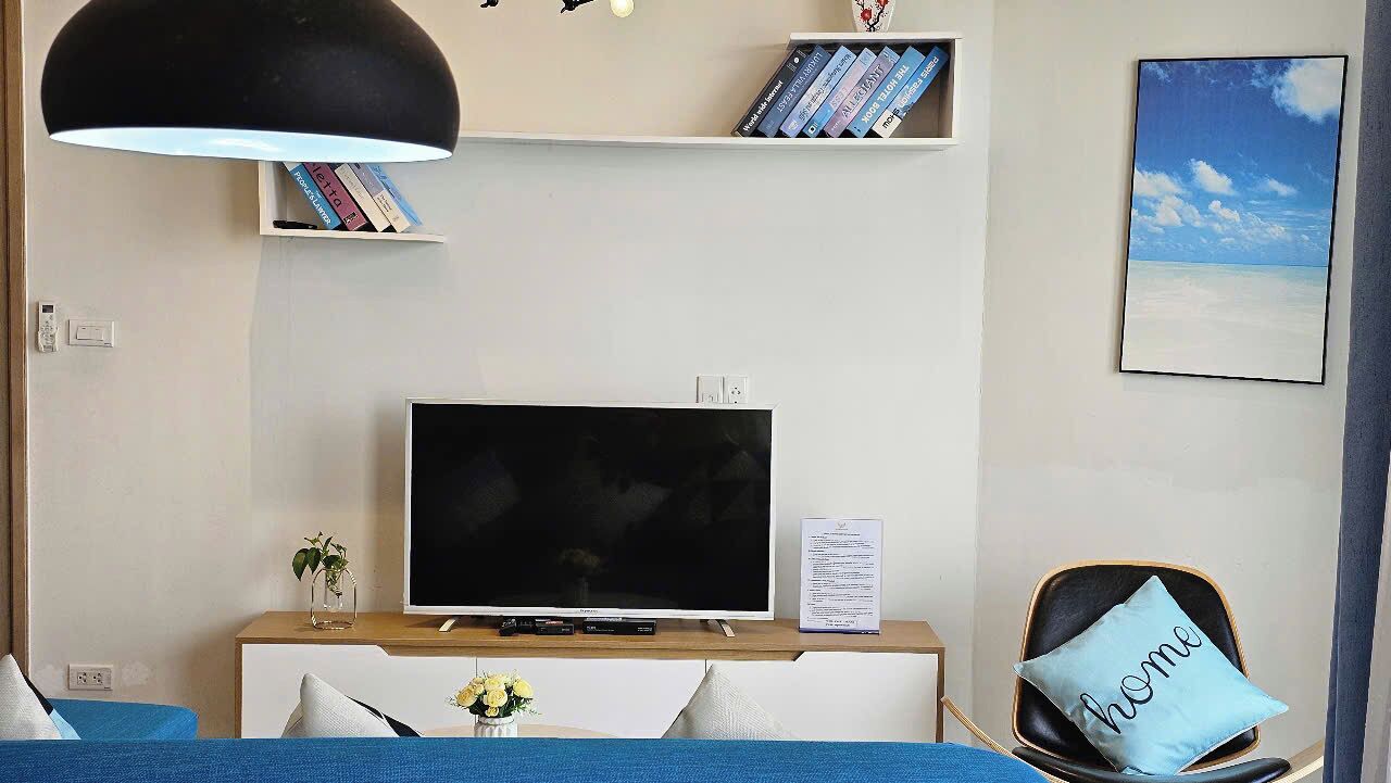 A modern hotel room with a flat-screen TV, a bookshelf with books, a framed picture on the wall, and a chair with a pillow.