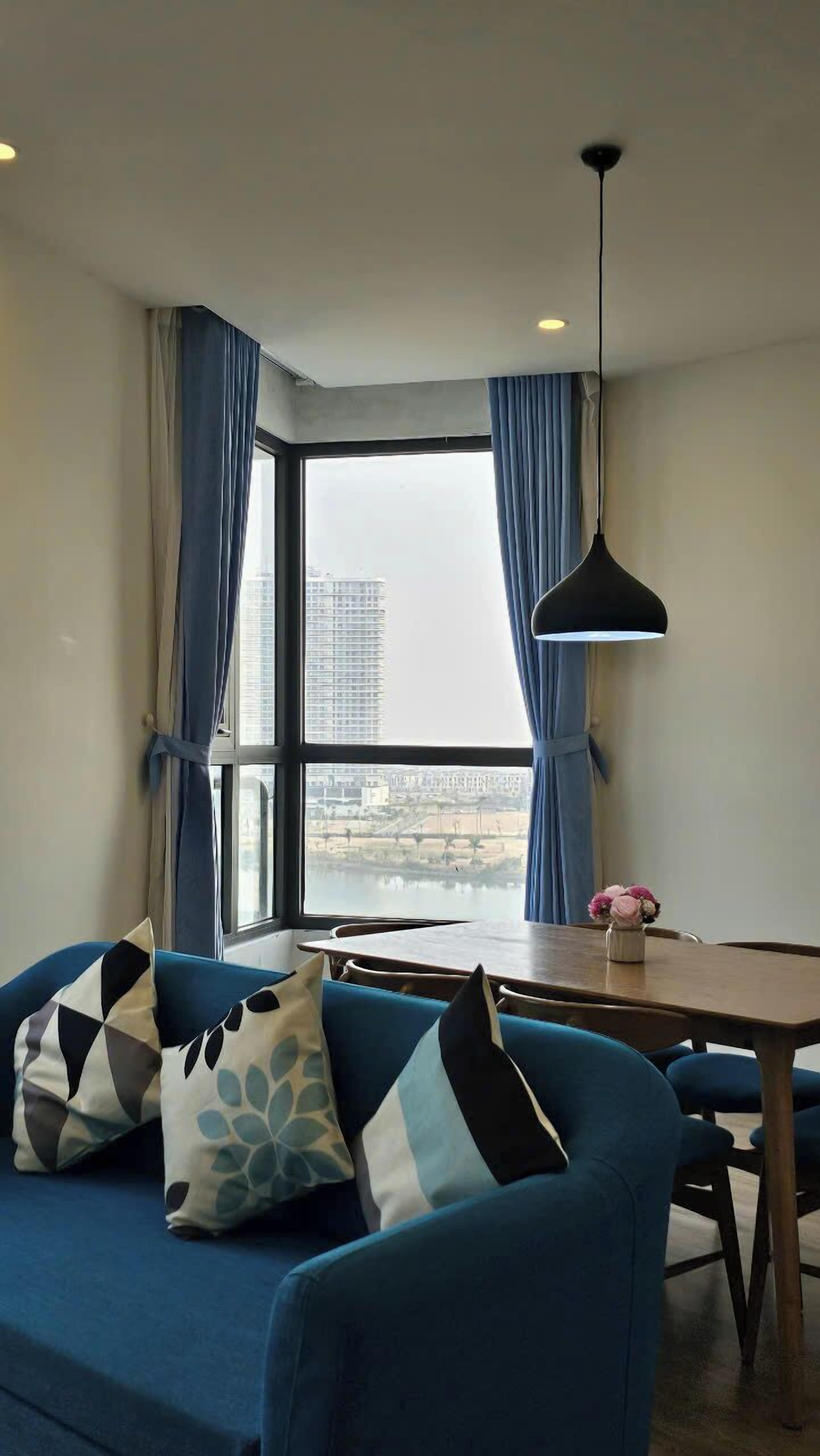 A modern living room with a blue sofa, a wooden dining table, and a view of a cityscape through large windows with blue curtains.