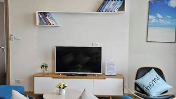 En moderne stue med et fladskærms-tv på et træstel, en reol med bøger, en lille plante og en 'home'-pude på en sort stol.