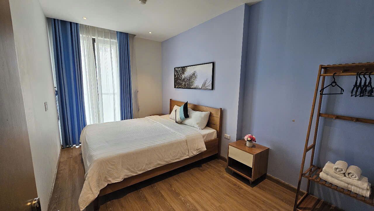 Un dormitorio con una cama, una mesita de noche, un estante en la pared con toallas y una ventana con cortinas.