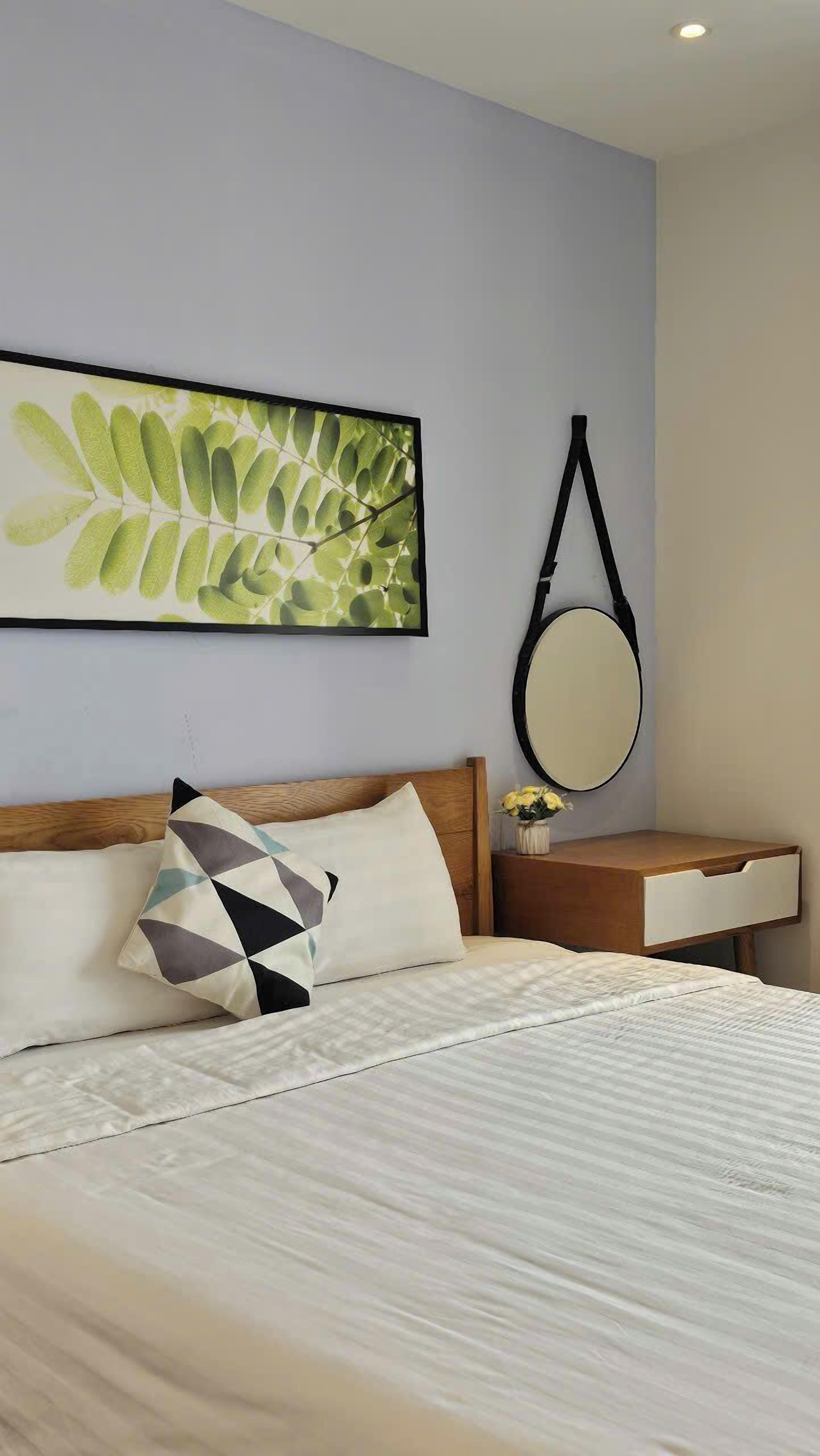 A neatly made bed with a patterned pillow, a bedside table with a mirror, and a wall-mounted picture of green leaves.