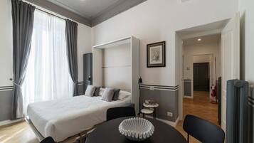 Ein modernes Hotelzimmer mit einem Bett, einem runden Tisch mit dekorativer Schale, einem kleinen Beistelltisch und einem Sessel. An der Wand hängt ein gerahmtes Bild, und das Zimmer ist in neutralen Farben gehalten.
