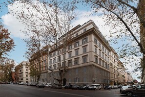 Un edificio de varios pisos con arquitectura clásica, que presenta una entrada en esquina y una fila de coches estacionados a lo largo de la calle.