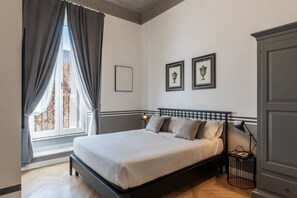 A bedroom with a bed, a window with curtains, a bedside table with a lamp, and framed pictures on the wall. - Castro Pretorio 24 - FromHometoRome (Roma)