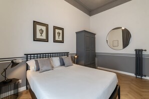 Ein Schlafzimmer mit einem Bett, zwei gerahmten Bildern, einem Kleiderschrank, einem runden Spiegel, einem Heizkörper und einem Nachttisch mit einer Lampe.