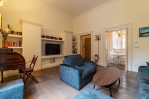 Ein Wohnzimmer mit blauem Sofa, einem Holztisch und einem Fernseher an einer weißen Wand. Es gibt Bücherregale und einen Kamin.