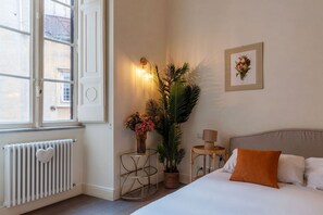 A bedroom with a bed, a radiator, a window, a wall-mounted light fixture, a framed picture, and a potted plant.