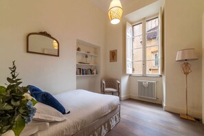 Een slaapkamer met een bed, een stoel, een radiator, een boekenplank en een raam met uitzicht op gebouwen.