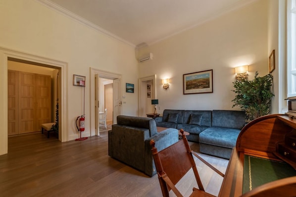 Ein geräumiges Wohnzimmer mit grauem Sofa, einem Holzstuhl und einem kleinen Tisch. Rechts steht eine Pflanze und an der Wand hängt ein Bild.