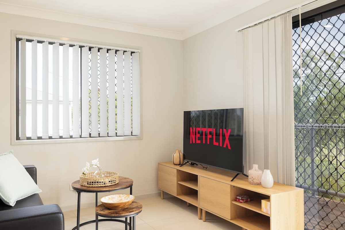 Een moderne woonkamer met een grote flatscreen televisie, een houten tv-meubel, een klein rond tafeltje met een mand en schaal, een bank en een raam met verticale jaloezieën.