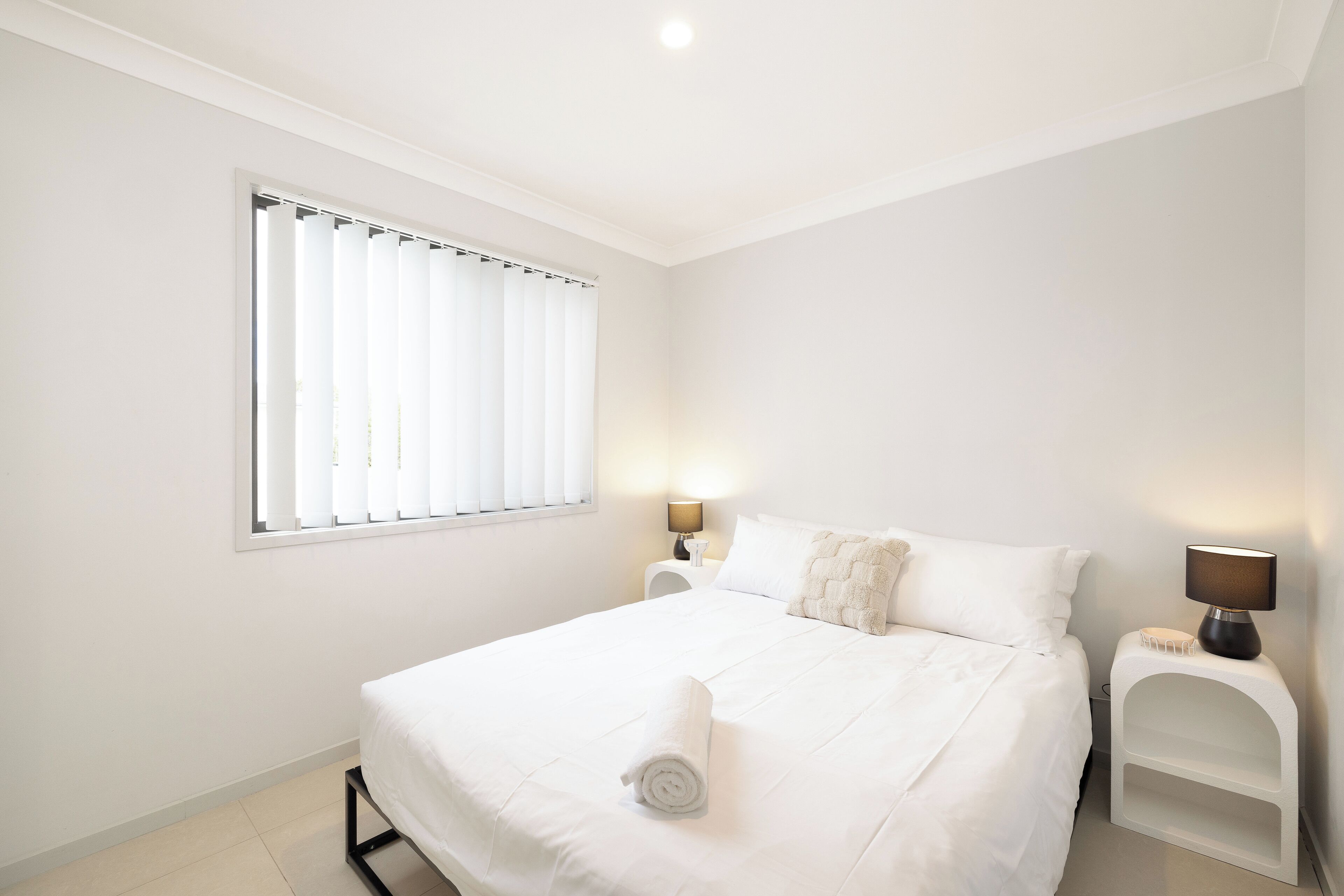 A white bedroom with a bed, a window with blinds, a bedside table with a lamp, and a small white shelf.