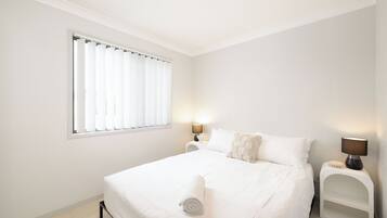 A white bedroom with a bed, a window with blinds, a bedside table with a lamp, and a small white shelf.