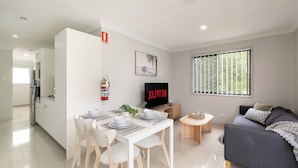 A modern living room with a grey sofa, a TV, and a dining area with a white table and chairs. - Smart Suites Self Check-in Apartments - 73 Bale (Rocklea)