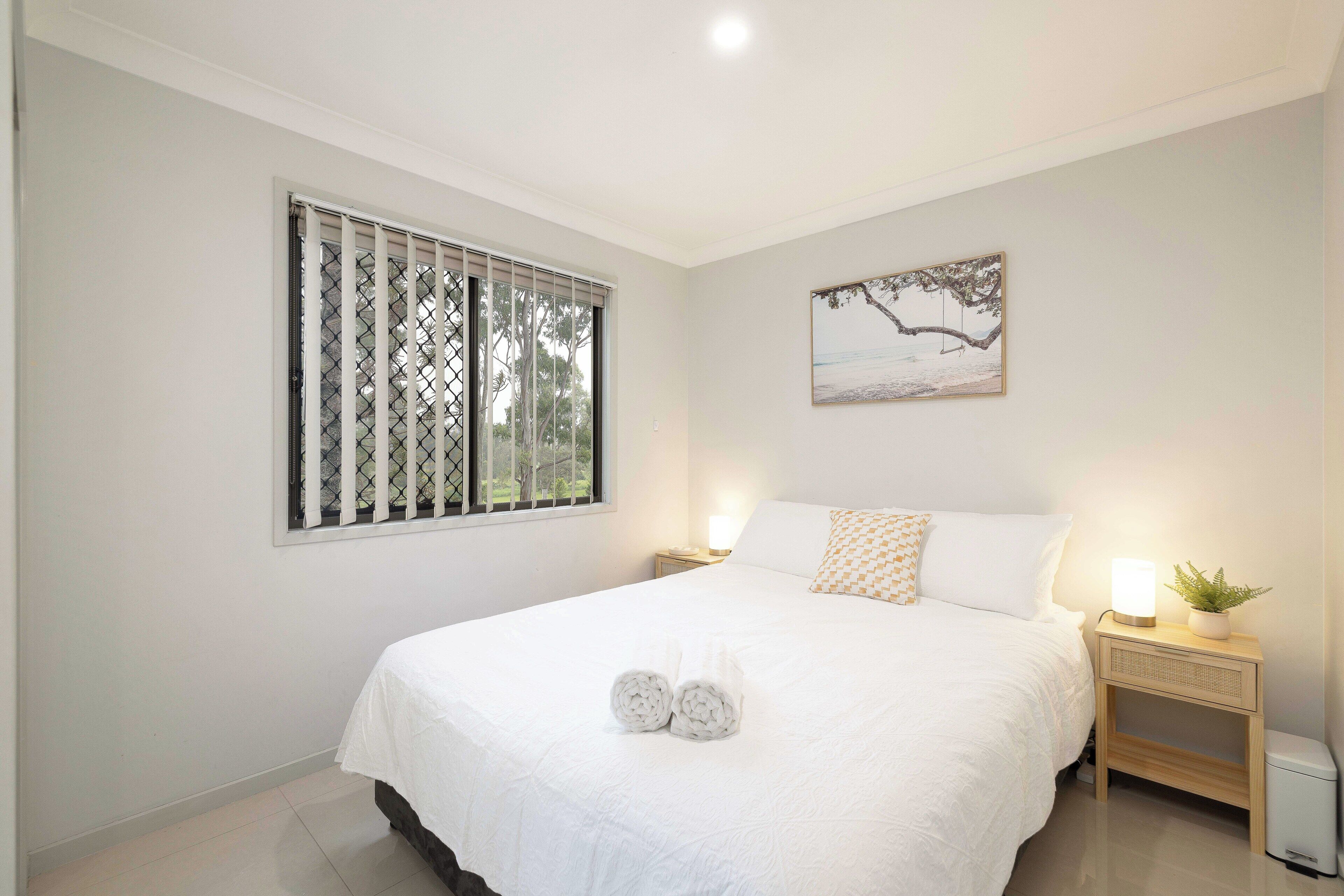 Un dormitorio con cama, ventana con persianas, mesita de noche con lámpara y un cuadro colgado en la pared.