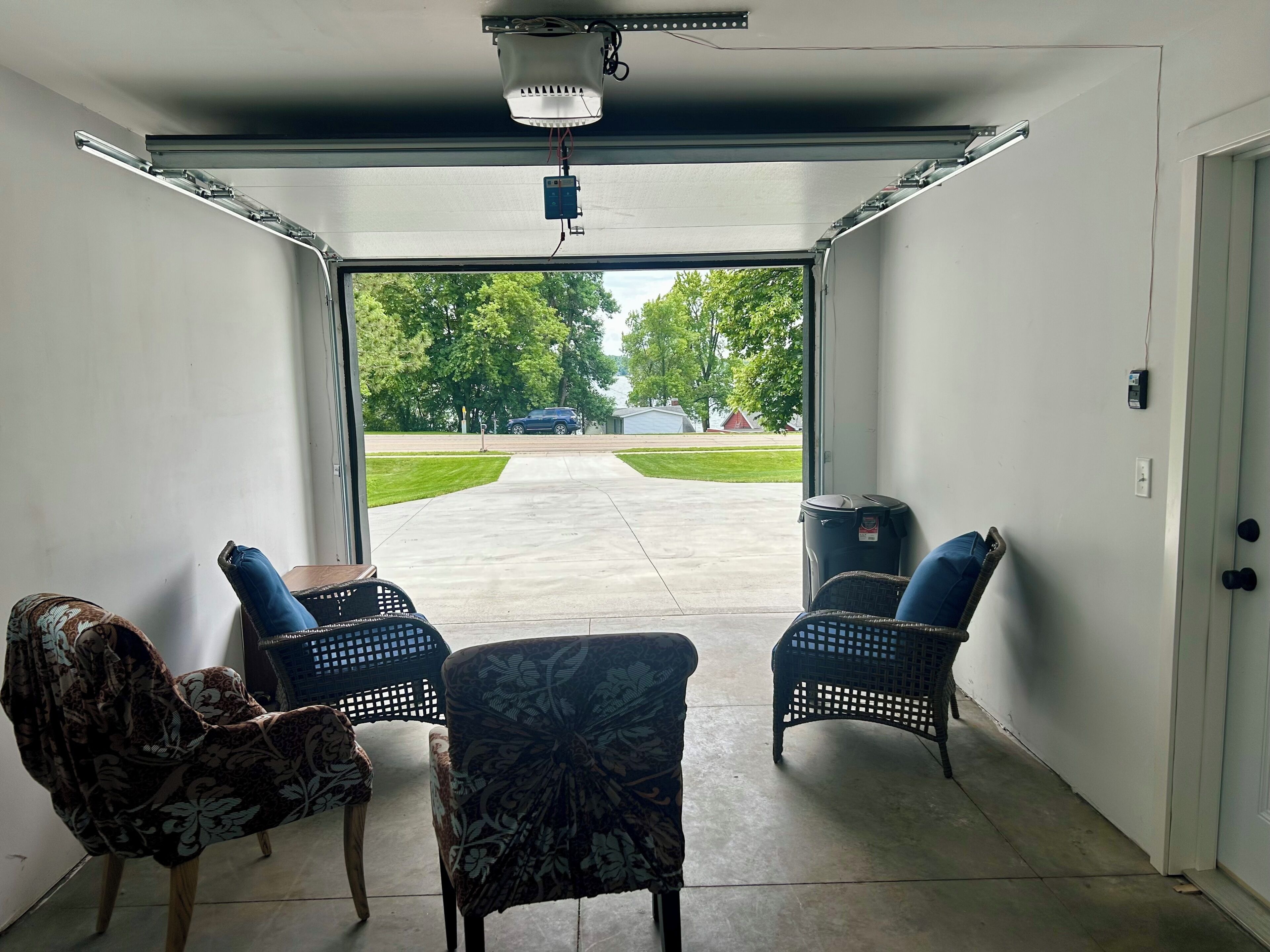 Een garage met twee rieten stoelen, een gestreept stoel, een plafondlamp en uitzicht op een oprit en bomen.