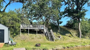 Un muelle de madera se extiende hacia un arroyo cristalino, una pequeña caseta con una puerta, y una escalera de madera que lleva a una terraza con un letrero que dice "The Trenchard's".