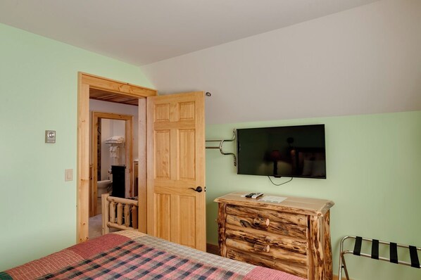 Un dormitorio con una cama, un aparador de madera, un televisor en la pared y una puerta que da al baño.