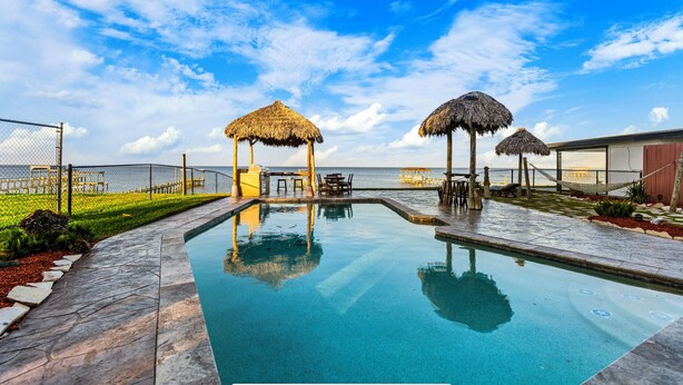 A swimming pool with two thatched-roof structures, a fenced area, and a view of the sea.