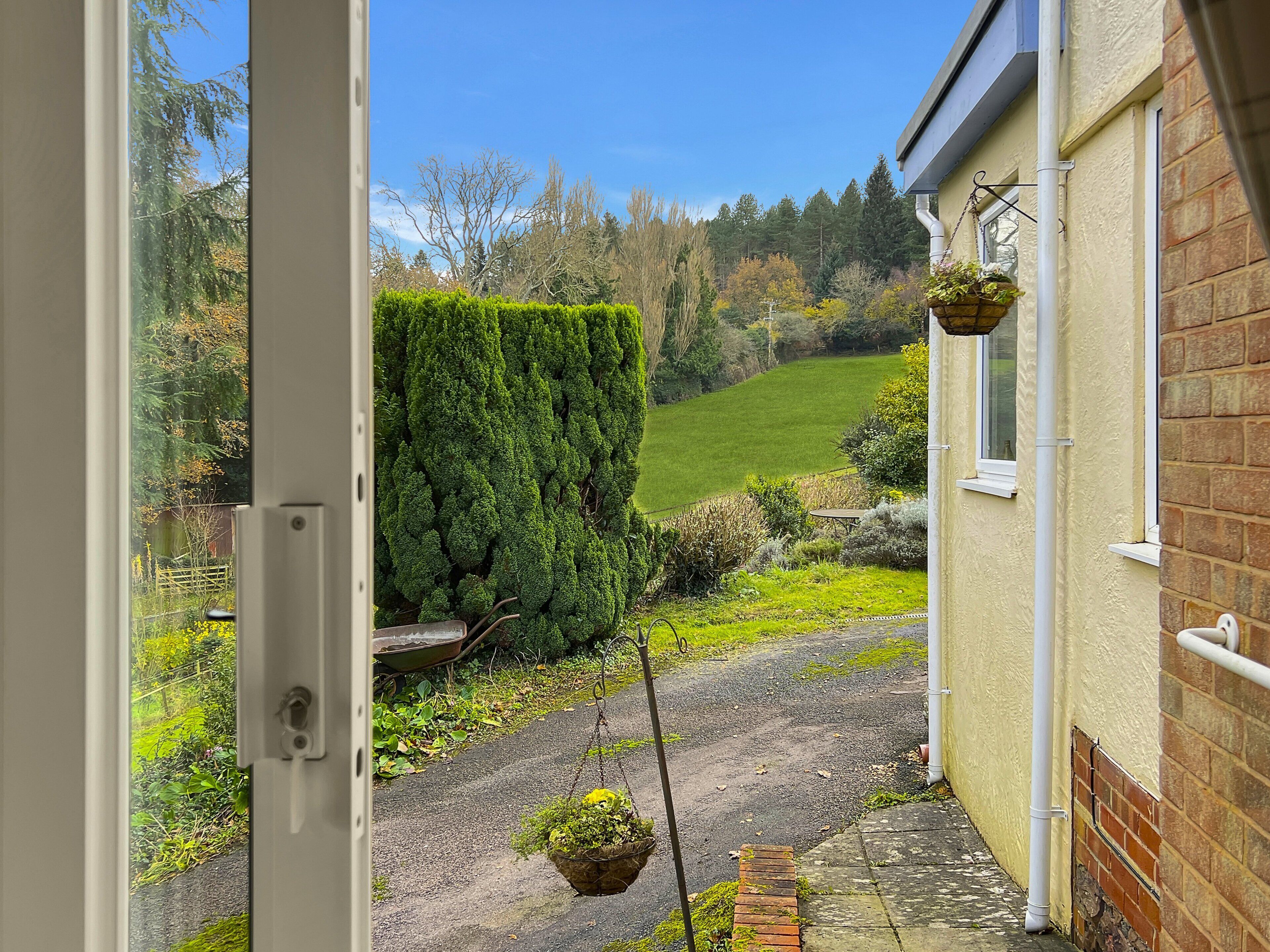 Une vue à travers la porte d’une maison, avec un chemin pavé menant à une pelouse verdoyante, une plante suspendue et un mur en briques.