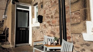 Un bâtiment rustique en pierre, doté d’une porte noire, d’un petit espace extérieur aménagé avec deux chaises en bois blanches et une table, ainsi que d’une boîte aux lettres noire fixée au mur.