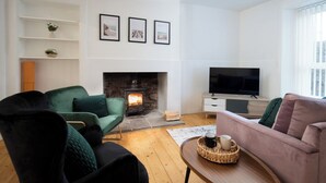 Un salon avec une cheminée, des fauteuils verts et noirs, un canapé rose, un téléviseur et des tableaux encadrés accrochés au mur.
