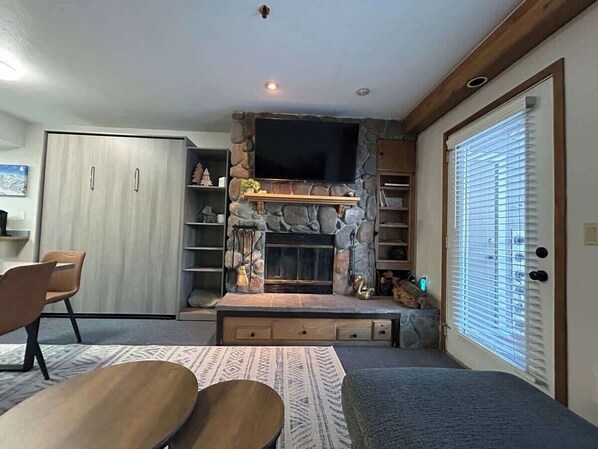 A living room with a stone fireplace, a television mounted above it, and a door with blinds.