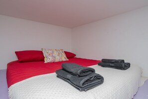 Ein Bett mit einer rot-weißen Tagesdecke, einem farbenfrohen Kissen und zwei gefalteten grauen Handtüchern.