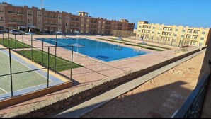 A swimming pool with a surrounding fence, a tennis court, and a building complex in the background. - Bayview Apartments Sahal Hasheesh  (Hurghada)